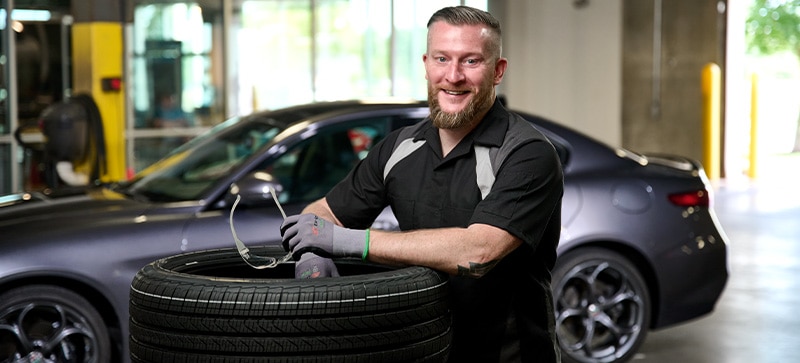 Recommended Installer Smiling Posed with a Tire Stack in an Auto Shop Garage